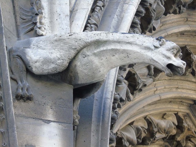 A long-necked, winged stone gargoyle jutting from the stonework beside the carved portal arches of Notre-Dame de Paris, in close-up.