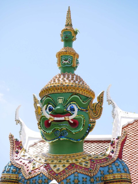 The head and shoulders of a green-faced giant guardian (yaksha) in mosaic armour at Wat Arun, Bangkok, against a pale sky.