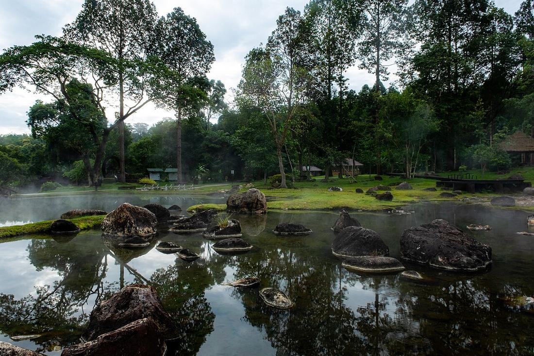 A natural hot spring pond surrounded by forest at Chae Son National Park in Lampang Province, Thailand. Steam rises from the mineral-rich water while rocks and trees are reflected on the calm surface.