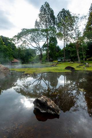 A natural hot spring pond surrounded by forest at Chae Son National Park in Lampang Province, Thailand. Steam rises from the mineral-rich water while the surrounding trees are reflected on the calm su