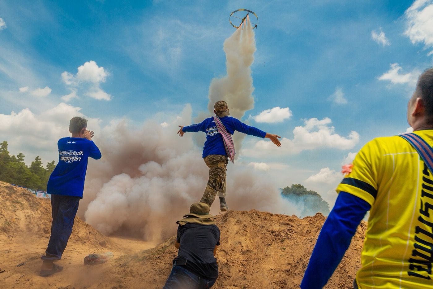 Men watch a bamboo rocket launch in a cloud of smoke against a blue sky